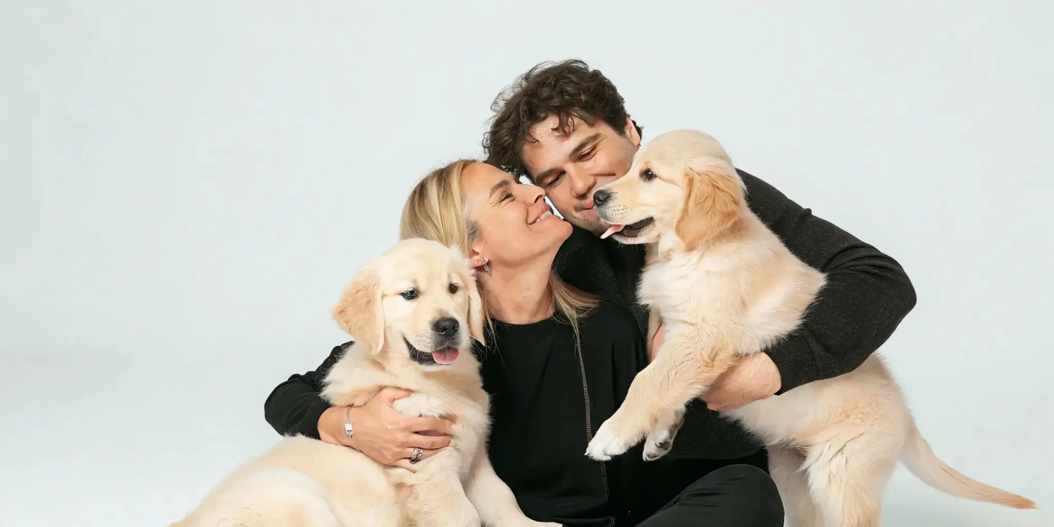 A couple lovingly holding two Golden Retriever puppies in a warm embrace.